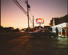 an old car parked in front of a do shop at sunset with people standing outside