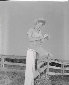 an old photo of a woman sitting on a fence post reading a book while wearing a hat