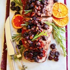 meat with oranges, olives and rosemary on a white platter next to utensils