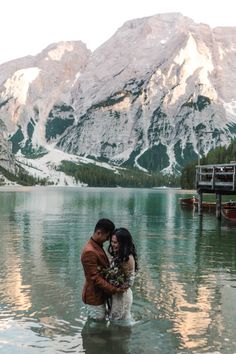 a man and woman standing in the water with their arms around each other near mountains