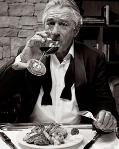 a man in a tuxedo drinking wine at a table with food and utensils