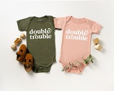 two baby onesuits that say double trouble and double trouble on them, next to shoes