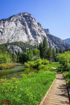 a wooden walkway leading to a river in front of a large mountain range with trees and grass