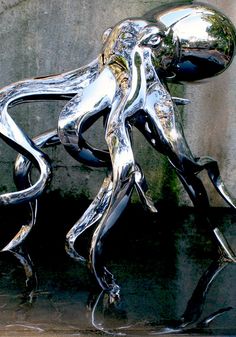 a shiny metal sculpture sitting on top of a wet ground next to a wall and water