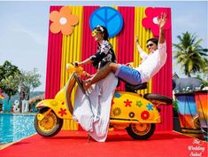 two people sitting on a motor scooter in front of a colorful wall and pool