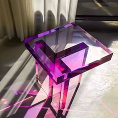 a purple glass table sitting on top of a floor next to a window with white curtains