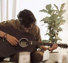 a woman sitting on the floor playing an acoustic guitar next to a potted plant