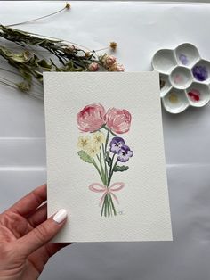 a person holding up a card with watercolor flowers on it, next to some other items