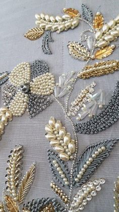 an embroidered piece with gold and silver beads on top of grey fabric, surrounded by leaves