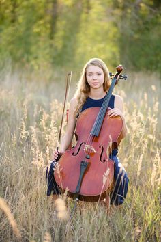 a woman holding a cello in a field with tall grass and trees in the background