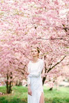 a woman wearing a white dress standing in front of trees with pink flowers on them