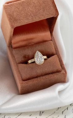 a gold ring with a pear shaped diamond sits in an open box on a white cloth