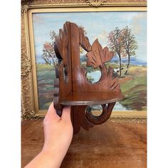 a hand holding a wooden clock in front of a painting on the wall with trees