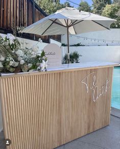 a wooden bar with an umbrella and flowers on it next to a swimming pool in the background