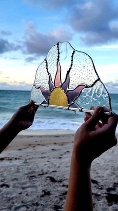 someone is holding up a stained glass flower on the beach with waves in the background