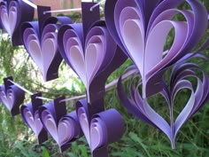 purple paper hearts hanging from clothes pins in front of trees and bushes with green grass