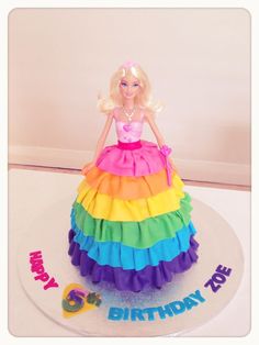 a birthday cake with a barbie doll sitting on it's top and rainbow ruffled skirt