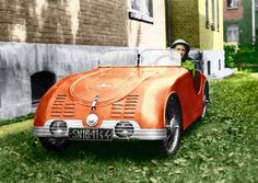 an orange car parked in the grass next to a brick building with a little boy sitting in it