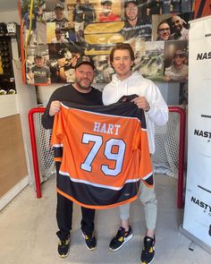 two men holding up an orange jersey in front of a wall with photos on it