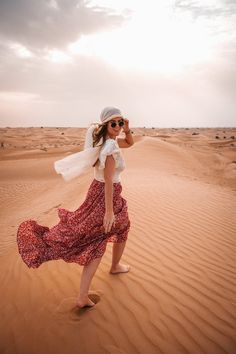 a woman standing in the desert wearing a white top and red floral dress with her arms around her head