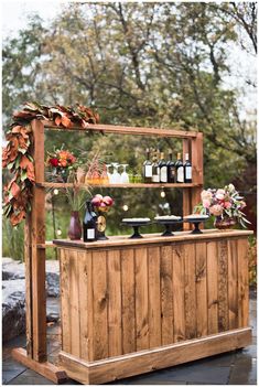 a wooden bar with bottles and flowers on it's top, next to a tree