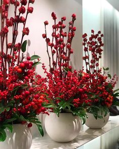 three white vases filled with red berries and greenery on top of a counter