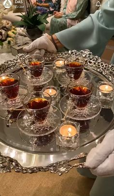 a tray filled with tea cups and saucers on top of a table next to people