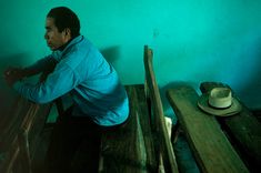 a man sitting on top of a wooden bench next to a green wall and hat