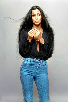 a woman with long black hair standing in front of a gray background wearing jeans and heels