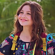 a woman with long brown hair wearing a colorful dress and smiling at the camera while standing in front of some trees