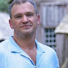 a man wearing a blue shirt and tie standing in front of a wooden structure with trees