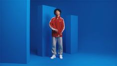 a man standing in front of a blue wall wearing a red jacket and white sneakers