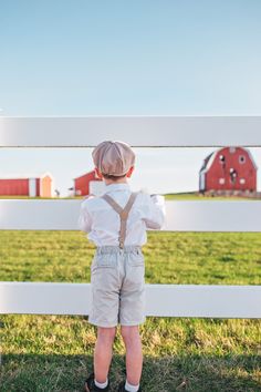 a little boy standing in front of a white fence looking at the grass and farm