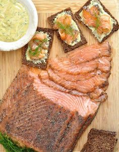 a wooden cutting board topped with slices of salmon and crackers next to a bowl of dip