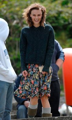 a woman in floral skirt and green sweater smiling at the camera while another man looks on