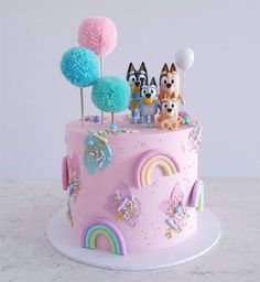 a pink cake decorated with cartoon characters and rainbow pom - poms on top