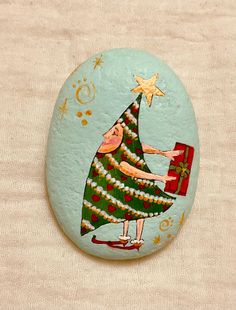 a painted rock with a christmas tree holding a present on it's side,