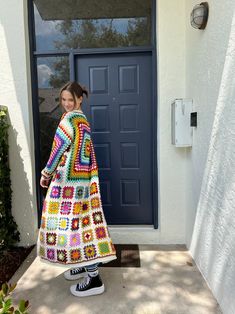 a woman standing in front of a blue door wearing a multicolored crocheted blanket