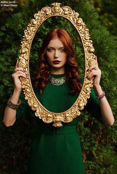 a woman with red hair holding up a gold framed mirror in front of her face