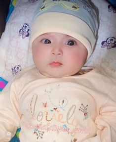 a baby laying on top of a bed wearing a white shirt and a blue hat