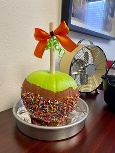 an apple decorated with sprinkles and a bow sitting on a desk next to a fan