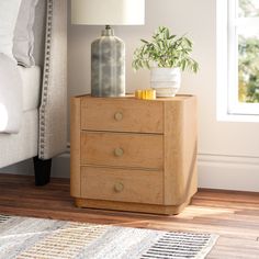 a nightstand with two drawers and a plant on it in front of a white bed