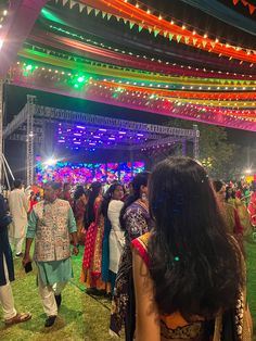 many people are standing around in front of an outdoor stage with lights on the ceiling