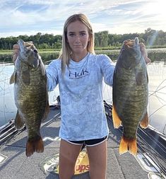 a woman holding two large fish in her hands on a boat with water and trees in the background