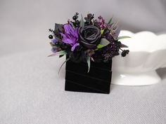 a black vase with purple flowers and greenery in it on a white table cloth