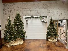 three small christmas trees in front of a white wall with bells on it and lights hanging from them