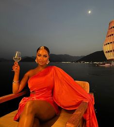 a woman in an orange dress sitting on a boat holding a glass of wine and looking at the camera