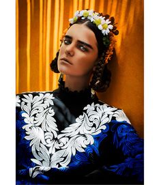 a woman with flowers in her hair wearing a blue and white dress sitting against a yellow curtain