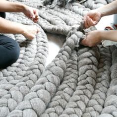 three people are sitting on a bed with a large chunky knitted blanket in the middle