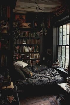 an unmade bed in a dark room with lots of books on the shelves and windows